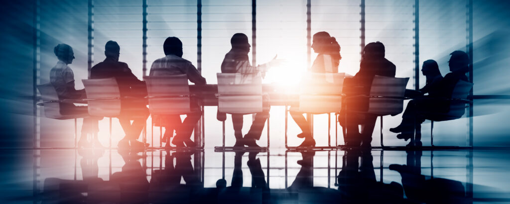 A group of business colleagues meeting at a long boardroom table with sunlight silhouetting them through the windows, representing the class action litigation services provided by the Toronto business litigation lawyers at Milosevic & Associates.