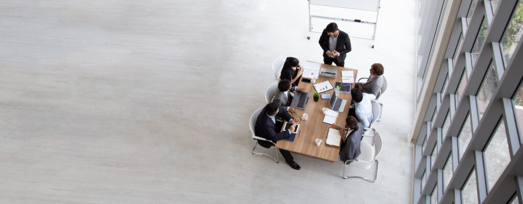 Aerial view of professionals meeting at a wooden conference table in a large, loft-style boardroom with a presentation board, representing the litigation services provided by Milosevic & Associates in Toronto in partnership disputes.