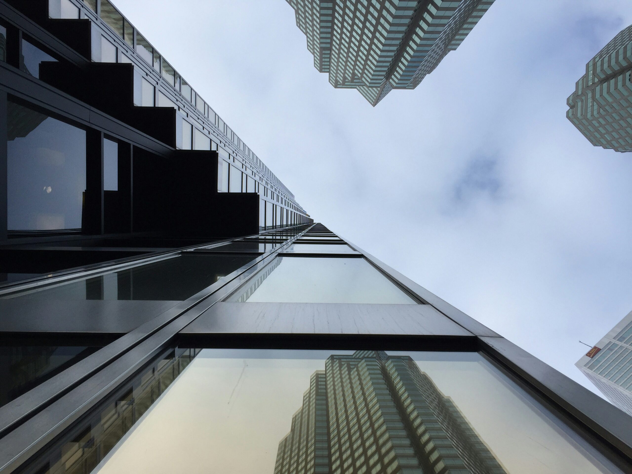 A three-point perspective image of a glass and black steel office building, stretching into a blue sky with white clouds, and one of the three nearby buildings is reflected in one of the window panes, representing derivative litigation.