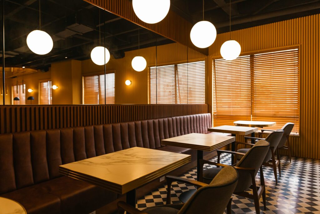A row of restaurant tables with faux marble tops and mirror walls over a continuous bench seat. Gray fabric and mid-century wood chairs are positioned opposite on a black and white checked floor, and the blind over the large window is shut, implying the business is closed, which represents fraudulent conveyance.
