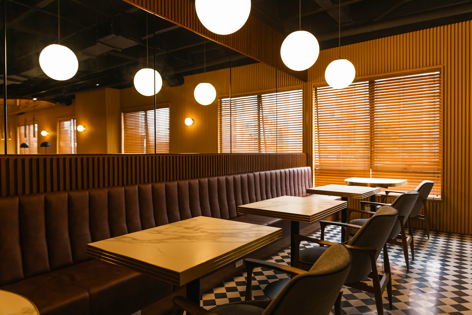 A row of restaurant tables with faux marble tops and mirror walls over a continuous bench seat. Gray fabric and mid-century wood chairs are positioned opposite on a black and white checked floor, and the blind over the large window is shut, implying the business is closed, which represents fraudulent conveyance.