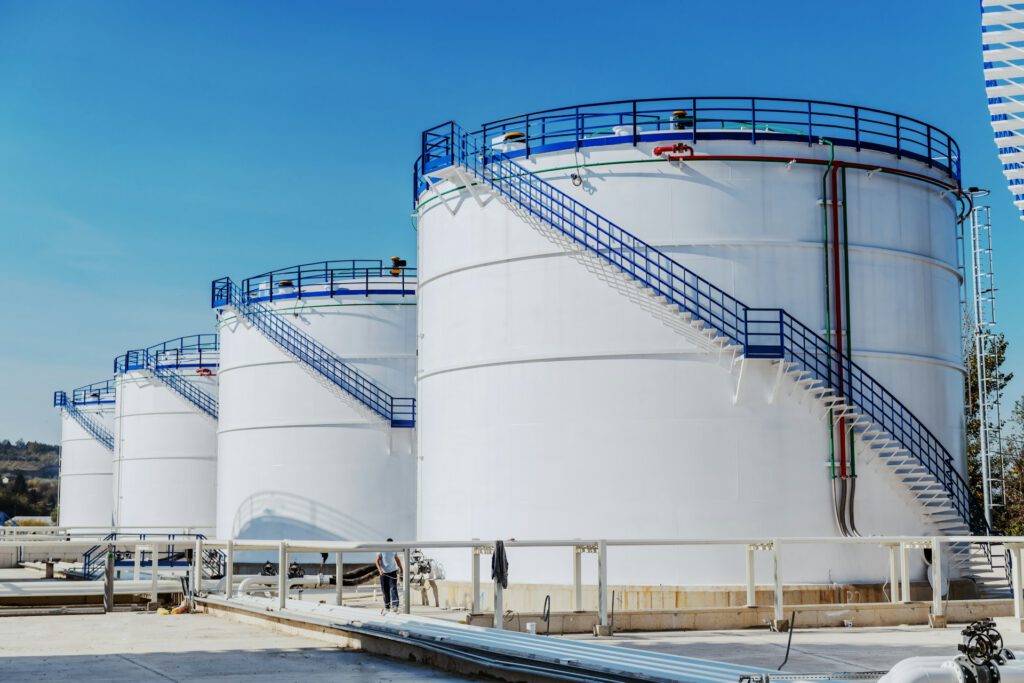 A row of oil tanks, each with stairs and blue railings curling around the exterior against a cloudless blue sky, representing Corporate Defamation.