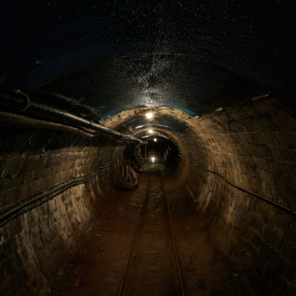 A mining tunnel with a metal track on the earth floor and cables and venting running the length of both curved brick walls, and intermittent bare bulbs glow from the black ceiling, similar to the mine discussed in this blog and representing Material Change Securities Act.