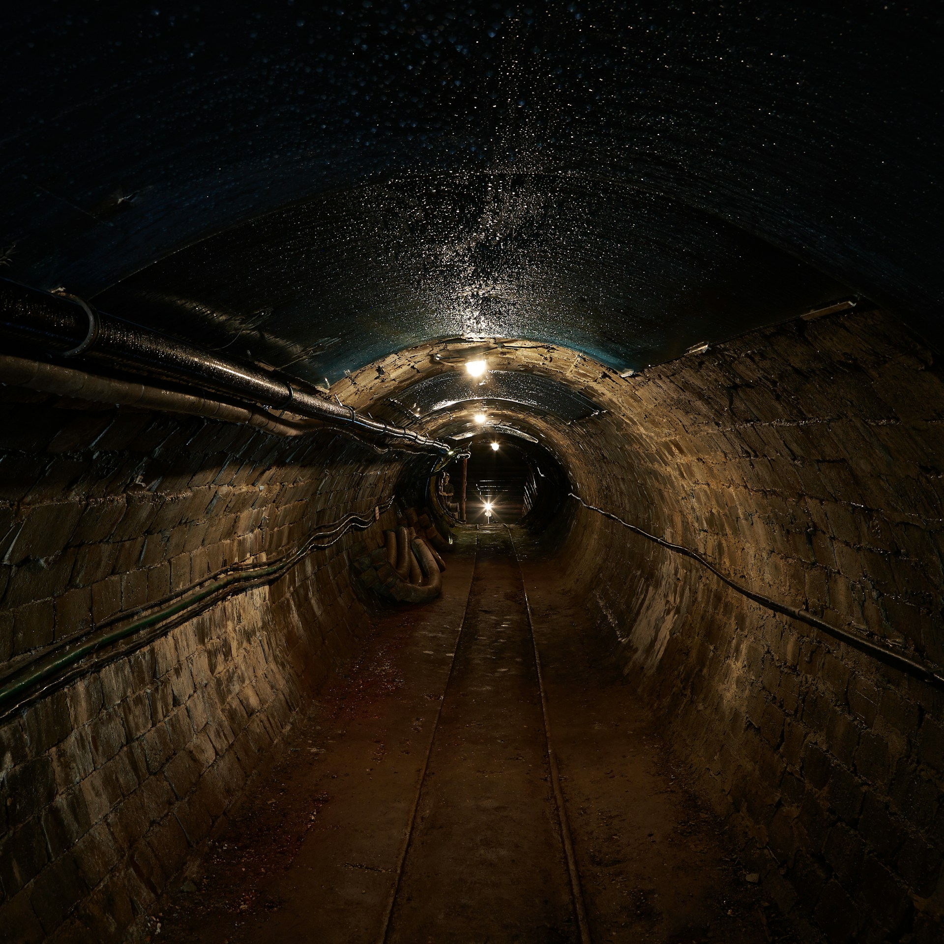 A mining tunnel with a metal track on the earth floor and cables and venting running the length of both curved brick walls, and intermittent bare bulbs glow from the black ceiling, similar to the mine discussed in this blog and representing Material Change Securities Act.