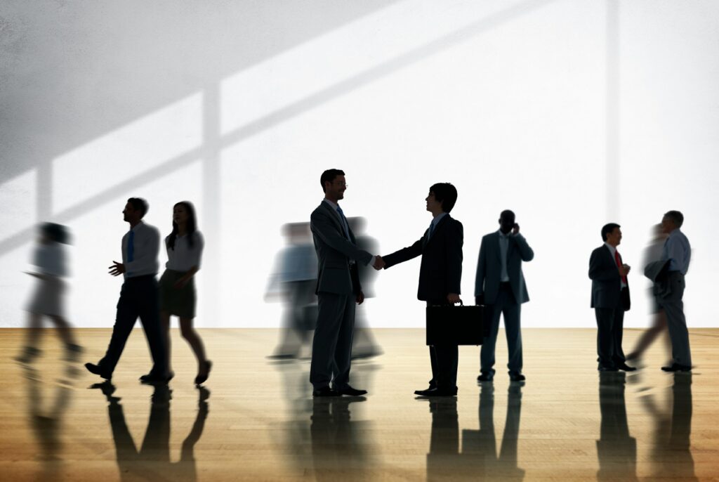Partially shadowed men in suits shake hands while other shadowed men and women walk and use phones in the background, representing David Milosevic OBA CPD chair.
