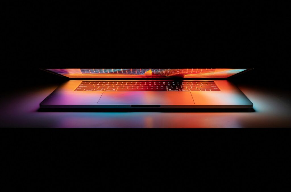 A partially open laptop, with orange, purple, and teal lights glowing from the screen and reflecting off the keyboard, is surrounded by darkness, representing defamation defences.