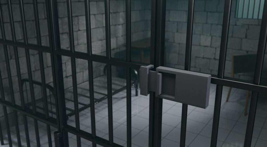 An empty jail cell with a chair, cot and not mattress, and gray brick walls through steel bars, representing Toronto investment fraud.