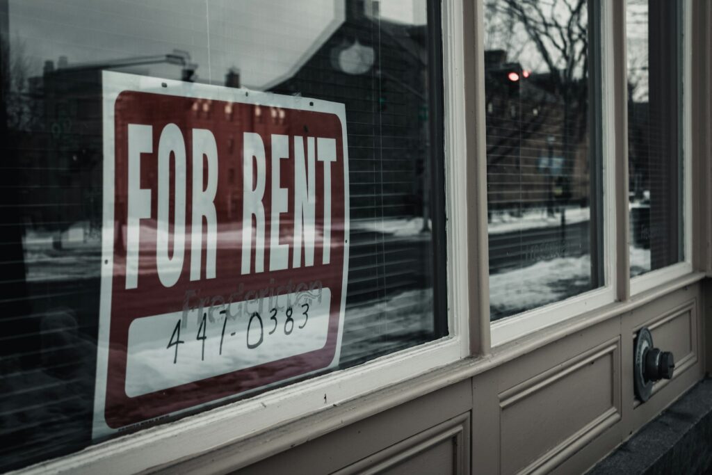 A For Rent sign is mounted in a window that reflects the houses on the opposite side of the street and the snow-lined road in the glass, representing rental fraud in Toronto.